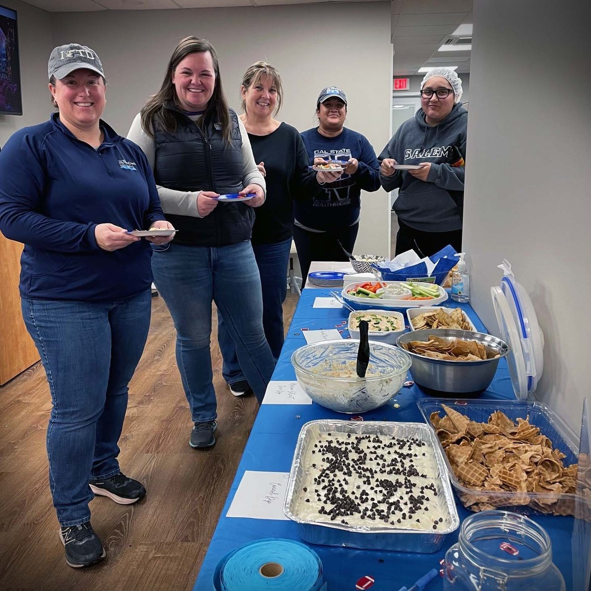 Five employees stand around a table with a blue table cloth. Multiple sweet and savory bowls of dips and chips or veggies sit on the table.