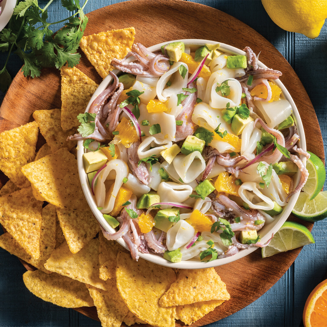 A round white dish briming with ceviche made of calamari rings and tentacles, orange slice, red onion, cilantro, and lime. Tortilla chips surround the dish.