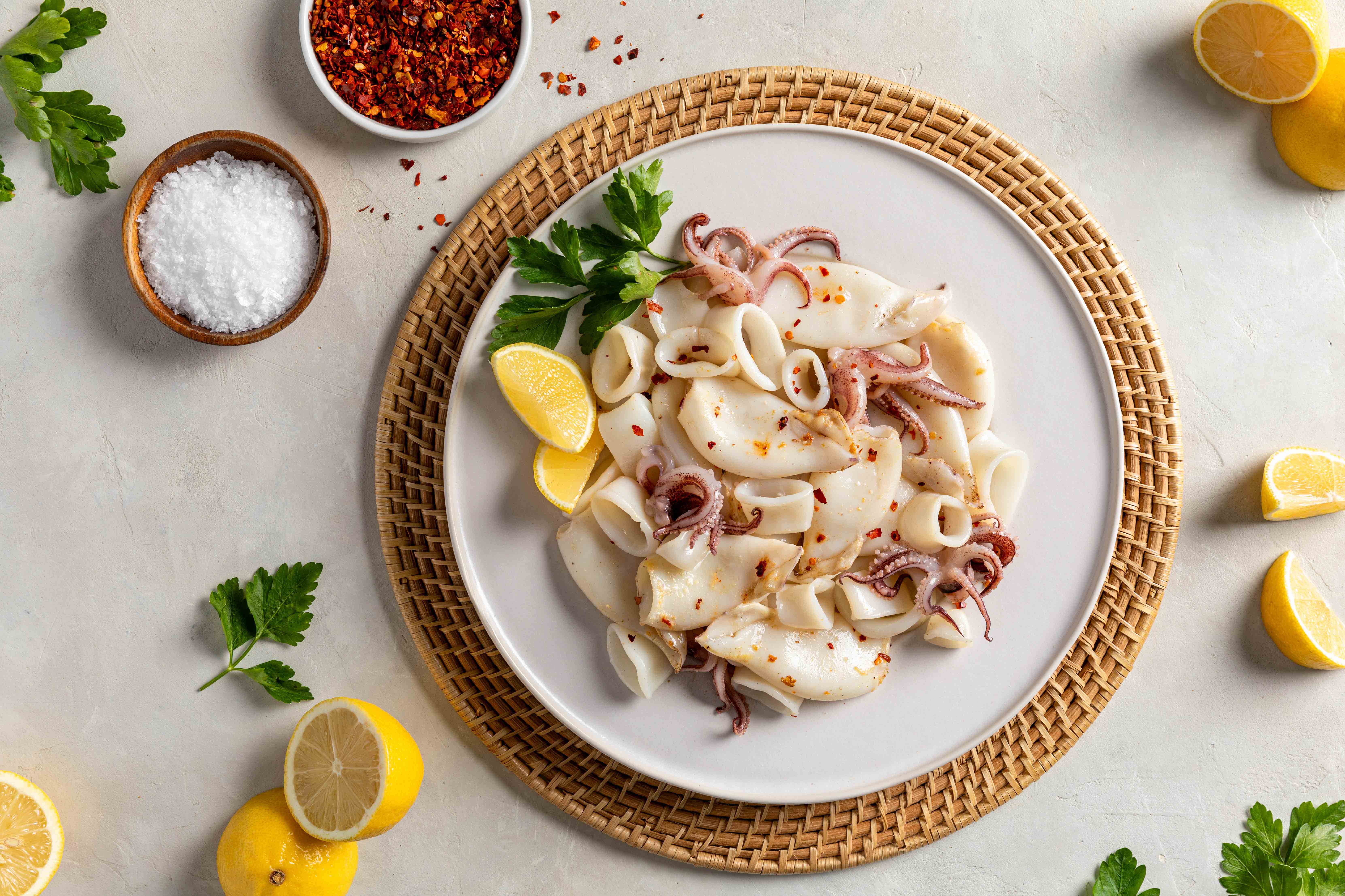 A white plate of calamari with traditionally cleaned (wings-on) tubes, tentacles, and rings. They are lightly cooked and have lemons and parsley.